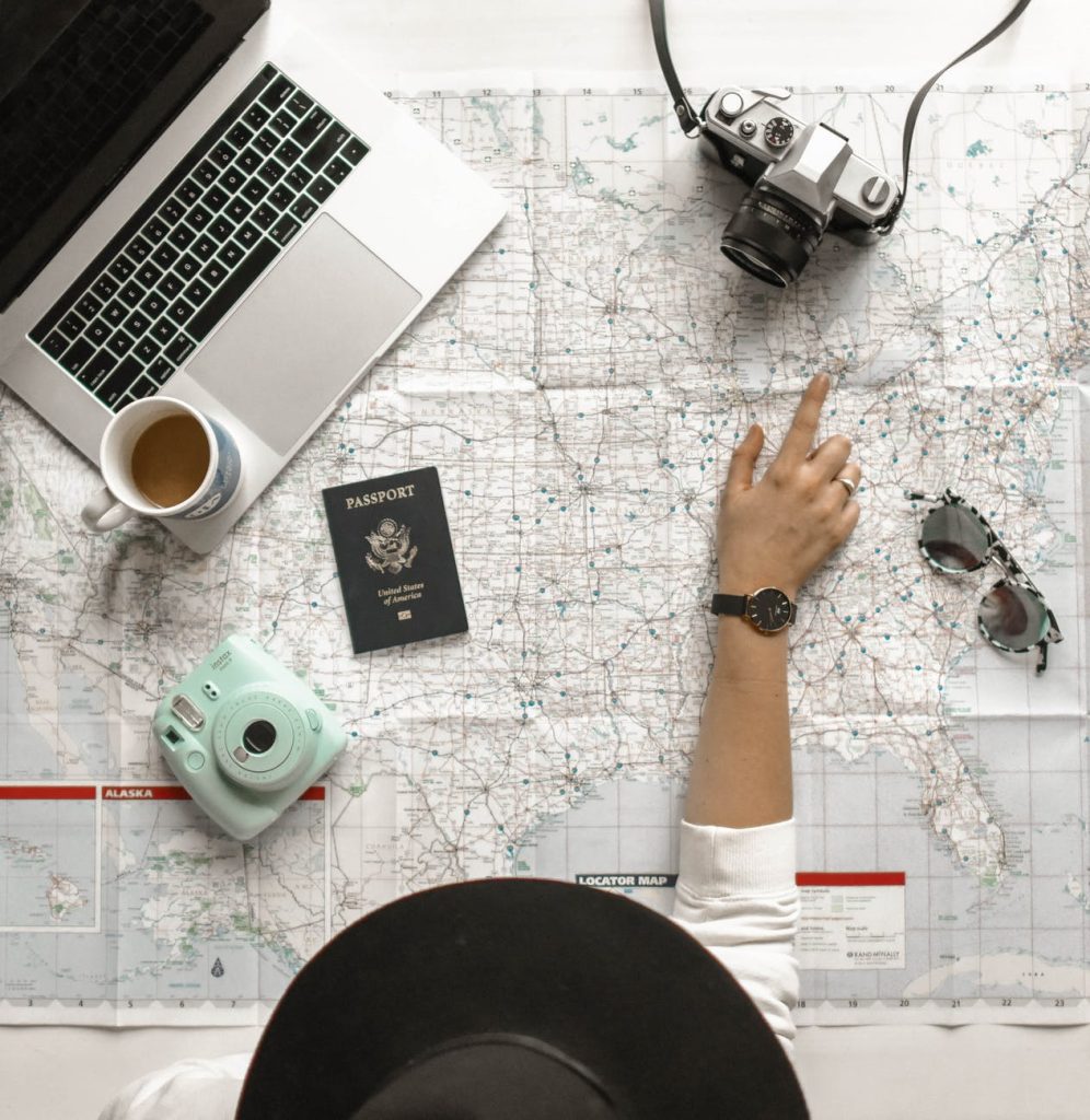 A person in a black hat points to a location on a map laid out on a table. Also on the table are a laptop, a cup of coffee, a camera, a green instant camera, a passport, and a pair of sunglasses. The scene suggests planning or reflecting on travel.