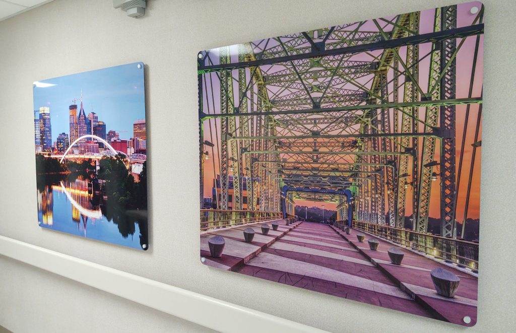 Photographs are displayed on a wall: the left image shows a city's skyline along a river at dusk, while the right image captures a view of a green steel bridge at sunset with purple, pink, and orange hues in the sky.