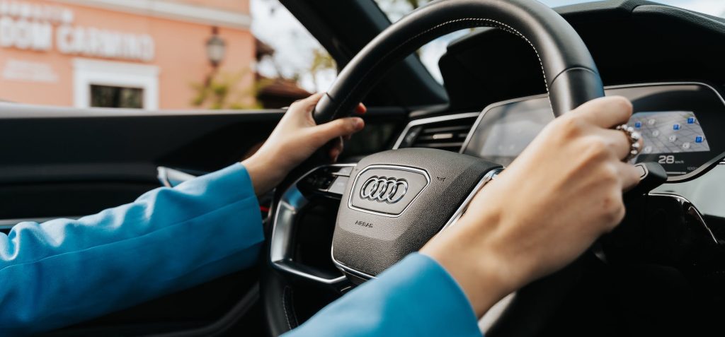 Close-up of a person’s hands holding the steering wheel of an Audi vehicle. The dashboard and digital display are visible, and the person is wearing a blue garment. The background shows an out-of-focus building.