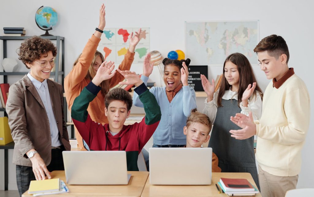 A group of six students, diverse in appearance, surround two laptops placed on a table in a classroom. They are raising their hands and cheering enthusiastically. An adult, possibly a teacher, stands nearby, smiling. The classroom has maps and globes as decoration.