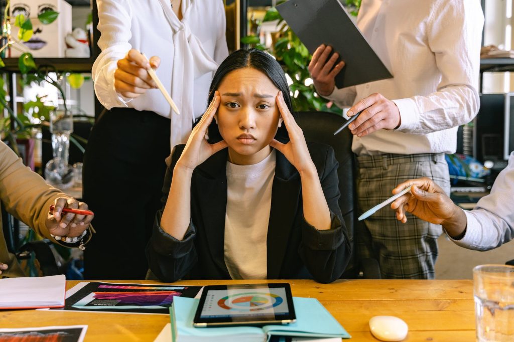 A stressed woman sits at a table full of documents and an iPad displaying a chart. She holds her head with both hands while multiple hands around her offer pens and papers, indicating pressure and demands. The background includes potted plants and office supplies.