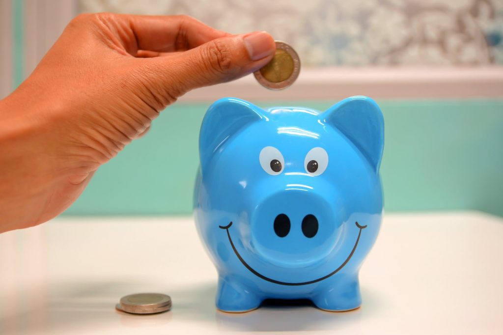 A person's hand is placing a coin into a blue piggy bank with a smiling face. There is another coin on a white surface next to the piggy bank. The background is blurred and features a light blue and white palette.