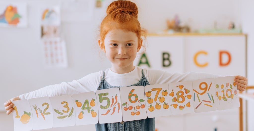 A young girl with red hair in a bun is holding a series of flashcards that display the numbers 1 to 10. Each number is illustrated with corresponding quantities of fruits and vegetables. The background shows a classroom with letters A, B, and C on the wall.