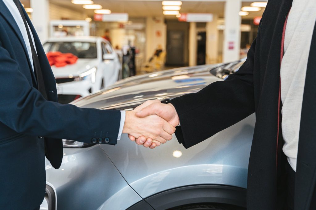Two people shaking hands in a car dealership, indicating a successful transaction or agreement. They are standing in front of a silver car with other cars visible in the well-lit showroom.