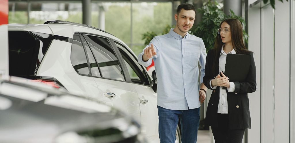 A man in a light blue shirt is discussing something with a woman in a black suit who is holding a black folder. They are standing in a car showroom, likely in conversation about a white vehicle that is partly visible in the foreground.