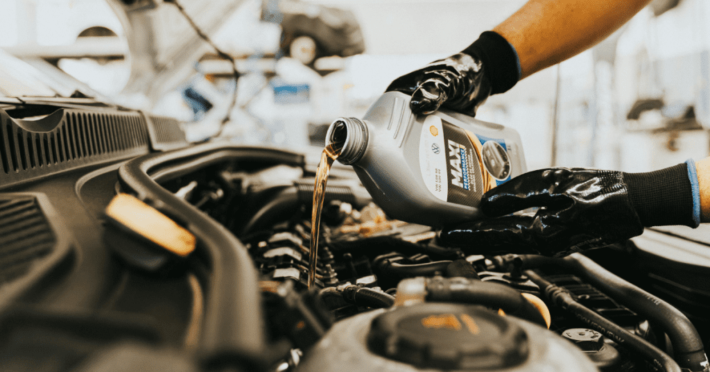 A person wearing black gloves pouring motor oil from a bottle into the engine of a car in a garage or workshop. Various automotive tools and equipment are visible in the background.