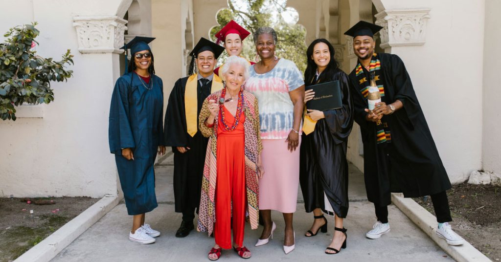 Seven people are standing and smiling outside. Three are wearing graduation caps and gowns, holding diplomas. An elderly woman in a red dress stands in the front, with an older woman and two others in casual clothes behind. They appear to be celebrating graduation.