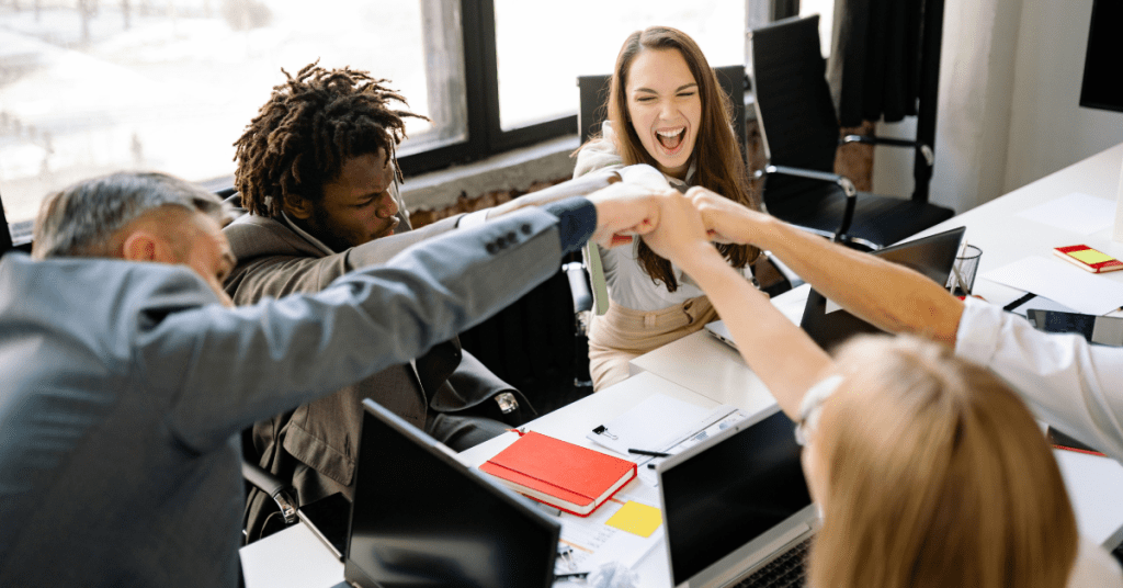 A diverse group of five office workers sitting around a table, smiling and cheering with hands outstretched toward the center for a fist bump. Laptops, notebooks, and office supplies are visible on the table, indicating a collaborative work environment.