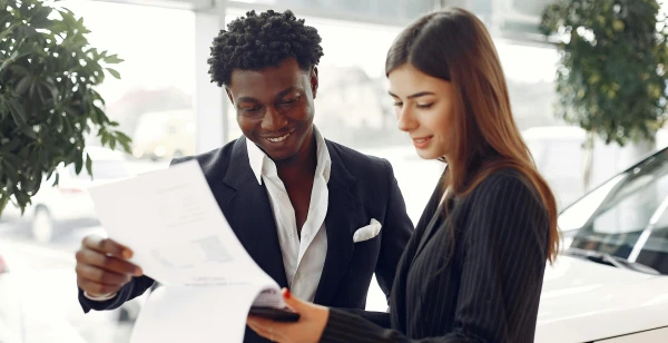 A man and a woman stand together in an office setting, discussing a project. The man, in a dark suit, is smiling and holding a document, while the woman, wearing a striped blazer, examines it attentively. Nearby, lush plants add vibrancy and there's even a sleek car parked outside.