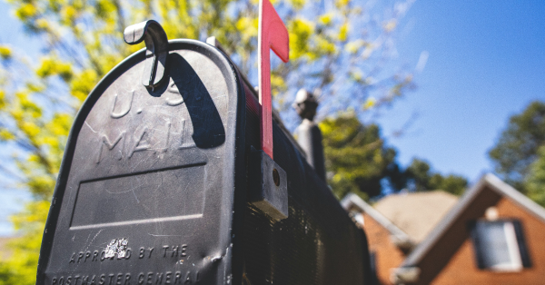 Close-up of a black U.S. mail mailbox with its signature red flag raised, akin to an auto draft notification, standing in front of a house. The background features a tree adorned with vibrant yellow leaves against a clear blue sky.