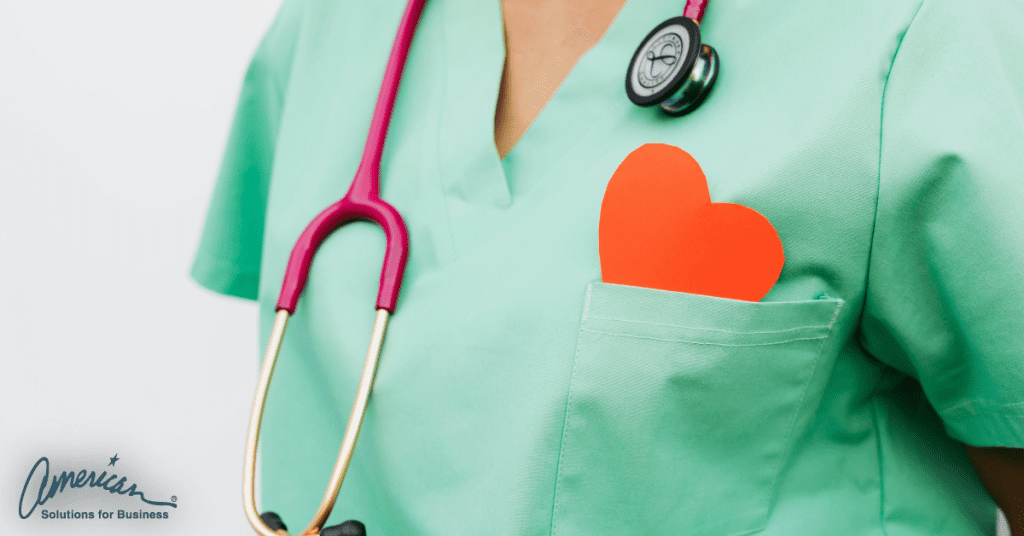 A person in green scrubs, with a pink stethoscope around their neck, carries a red paper heart partially tucked into their chest pocket. The 