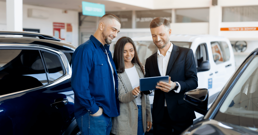 A smiling man and woman stand next to a car sales representative holding a tablet, discussing details inside a bright dealership with vehicles displayed around them—an ideal setting for engaging in-market buyers through combination marketing.