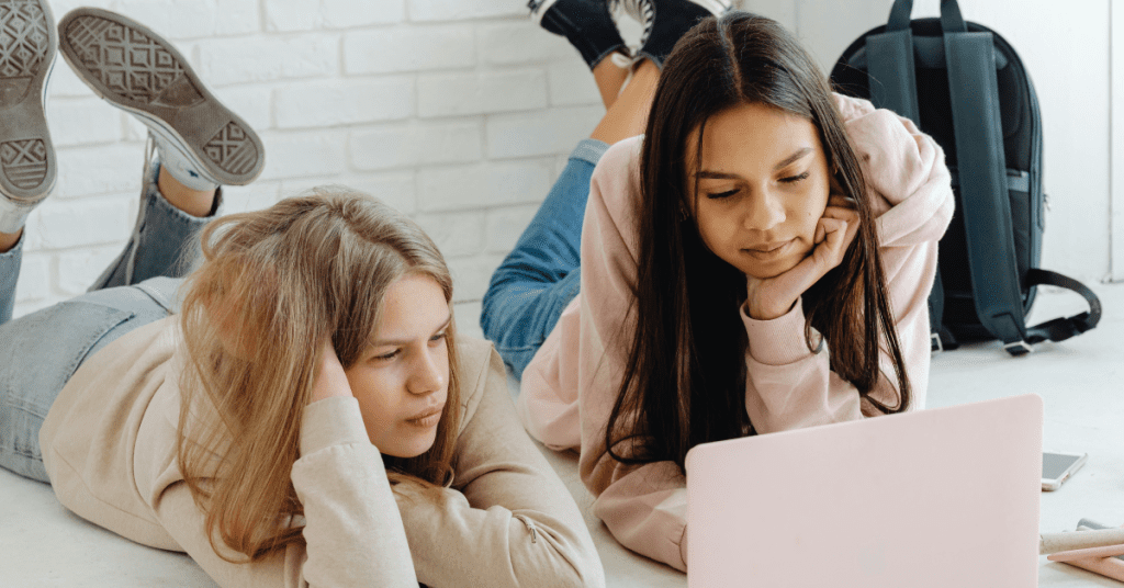 Two teenage girls are lying on the floor, looking at a pink laptop together. They appear focused and relaxed, possibly brainstorming fun fundraisers, with a black backpack and notebooks nearby against a white brick wall.