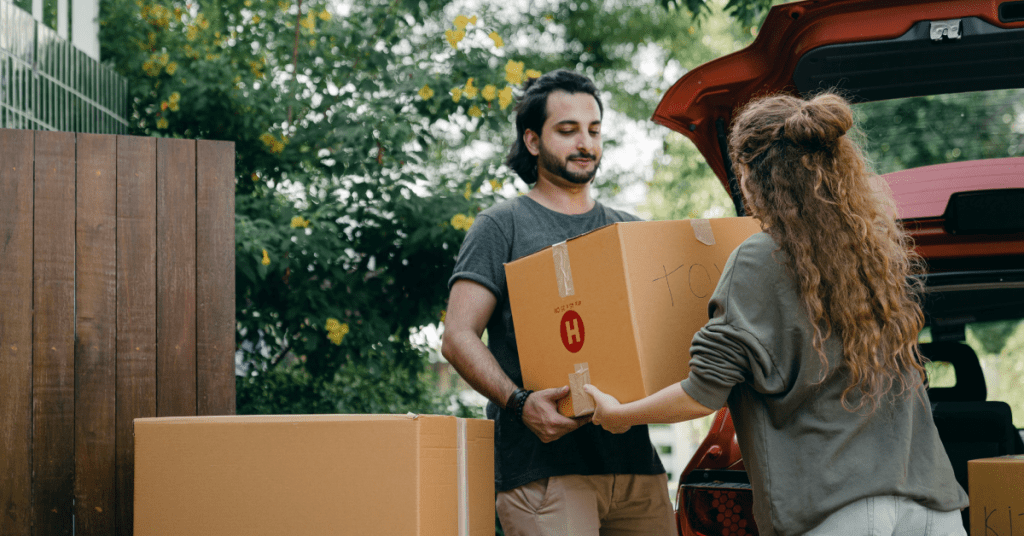 A man and woman unload cardboard boxes from the trunk of a red car outdoors, perhaps marking one of life’s big events. The man receives a box from the woman, with greenery and a wooden fence in the background.